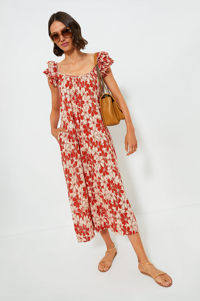 A woman wearing THE GREAT. red and white floral midi dress with ruffle sleeves, paired with tan sandals, a brown handbag, and sunglasses, stands against a plain white background.