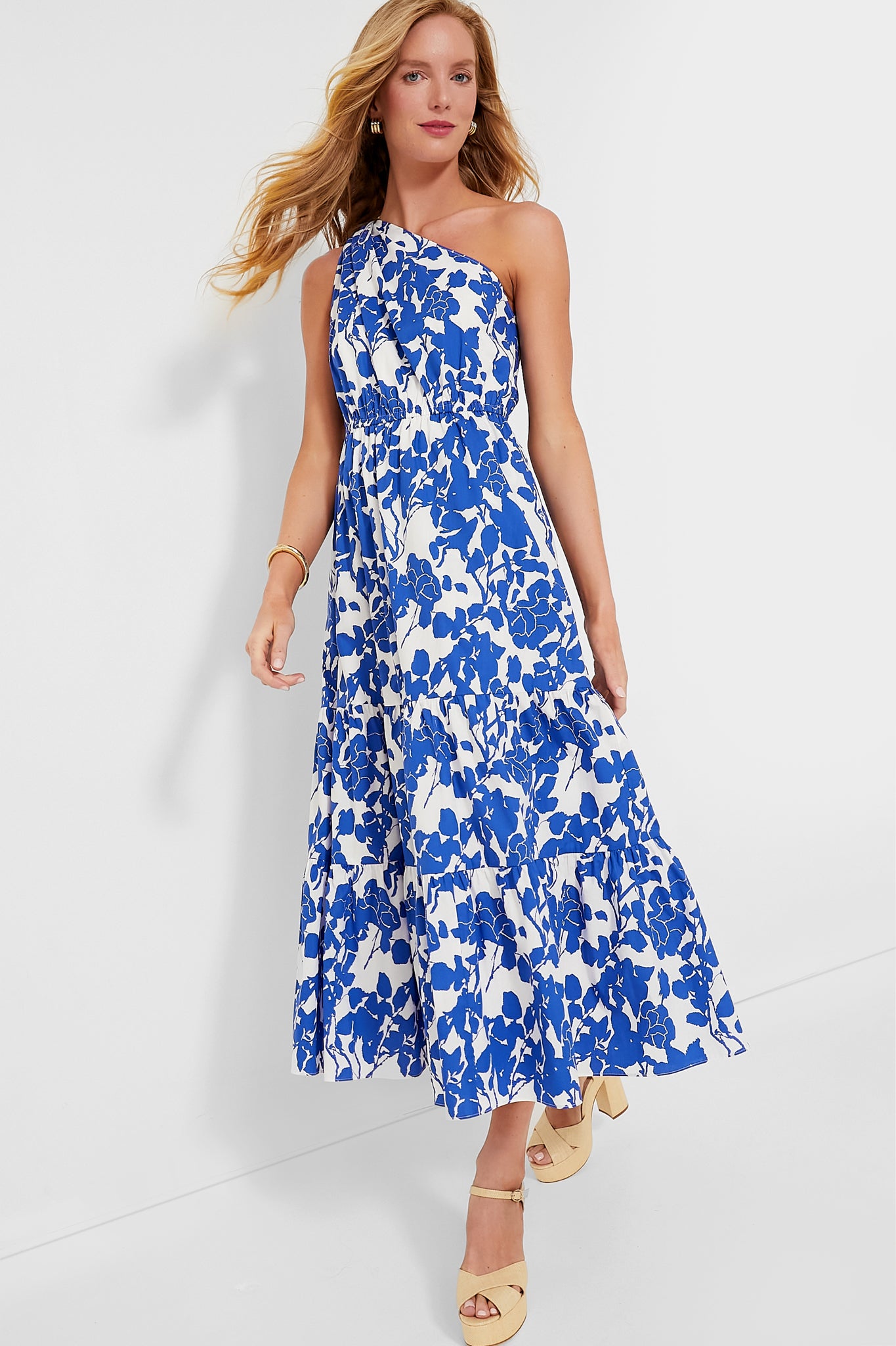A woman models a Hyacinth House blue and white cotton one-shoulder summer dress with a tiered skirt, paired with beige platform sandals, striking a stylish pose.