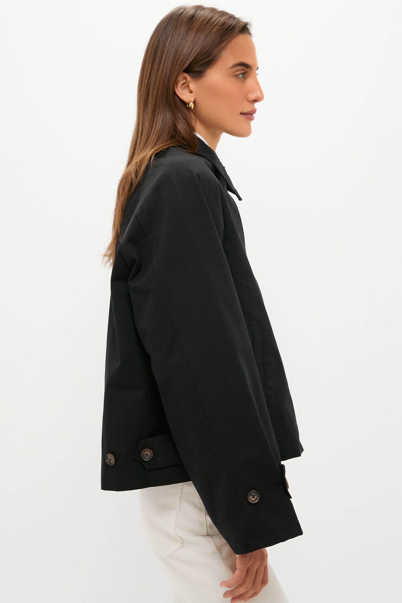 A woman stands in profile wearing a black cropped trench coat and white pants by Tuckernuck against a plain white background.