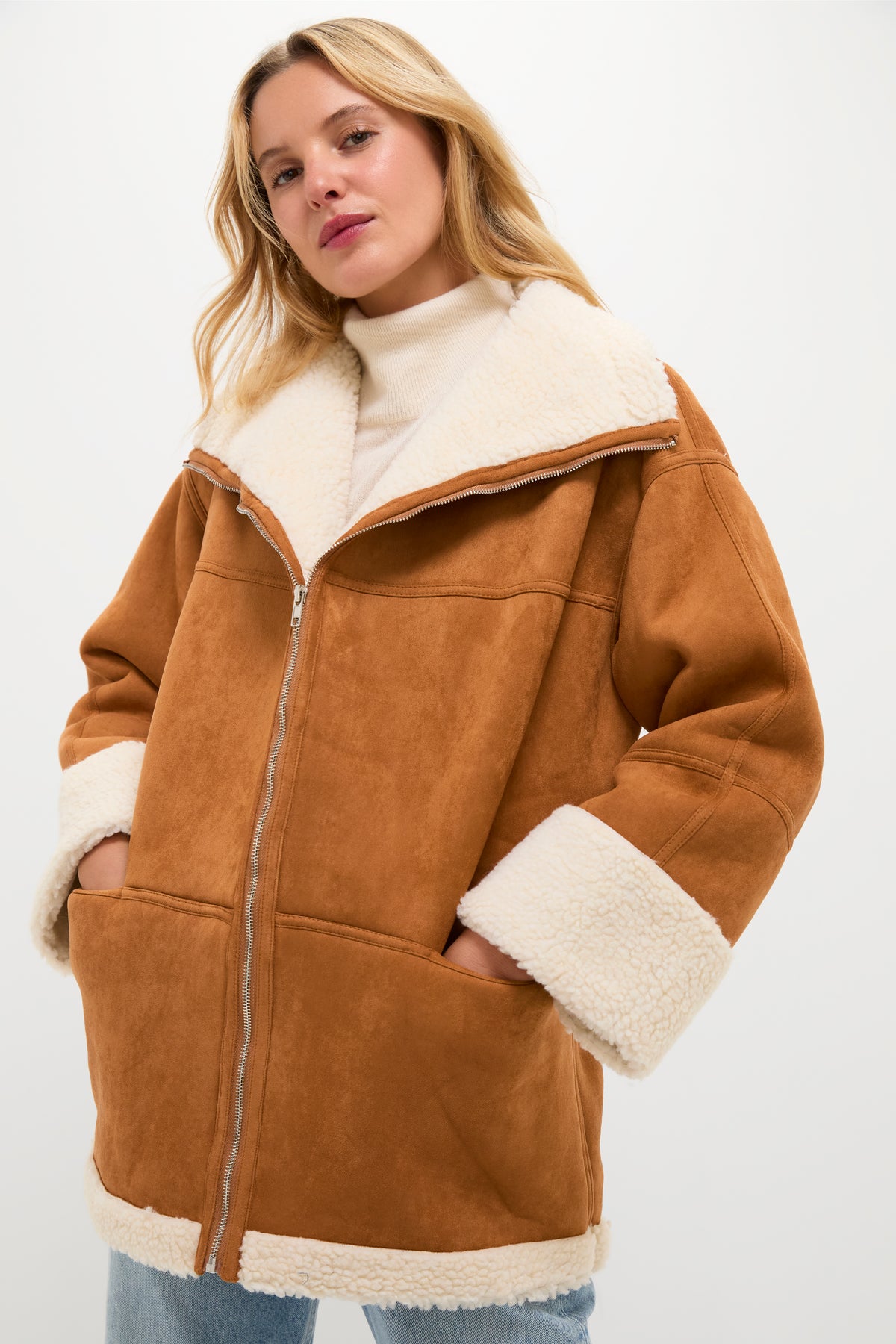 A person wears a Moon River camel suede coat with faux fur trim over a white turtleneck and blue jeans, hands in pockets, highlighting the oversized fit.