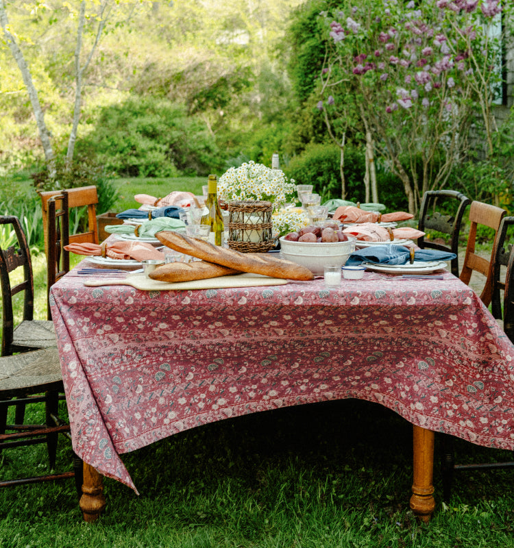 A table in the gardens set for an outdoor dinner. Table is set with a red tablecloth and surrounded by mix-matched chairs. 