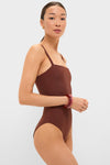Woman in profile wearing the classic Cappuccino Isabel One Piece swimsuit, featuring thin straps, styled with small gold hoops and red bangles, embodying effortless, high-end fashion.