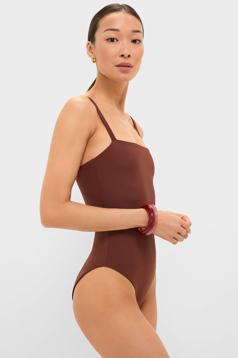 Woman in profile wearing the classic Cappuccino Isabel One Piece swimsuit, featuring thin straps, styled with small gold hoops and red bangles, embodying effortless, high-end fashion.