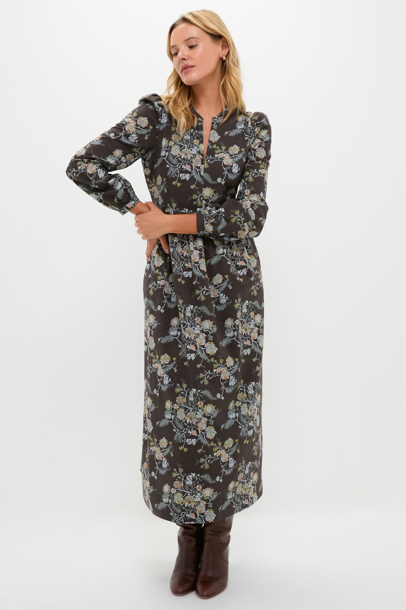 A woman stands against a plain background wearing a Hyacinth House dark floral maxi dress with blouson sleeves, a tie waist, and brown boots.