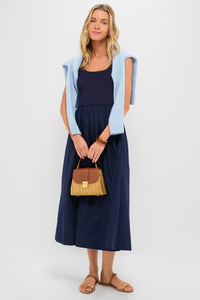 A woman wearing a navy Tuckernuck Juliette Dress with an A-line skirt, light blue sweater over her shoulders, small woven handbag in hand, and brown sandals.