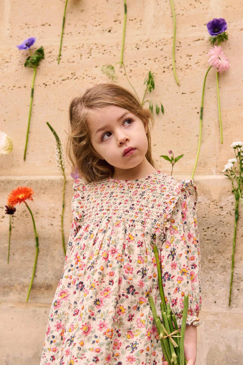 Young girl in a classic, high-end Cream Wild Flowers Saralou Dress, featuring smocked bodice and ruffle details, holding flower stems against a floral backdrop. Effortless and fun style.