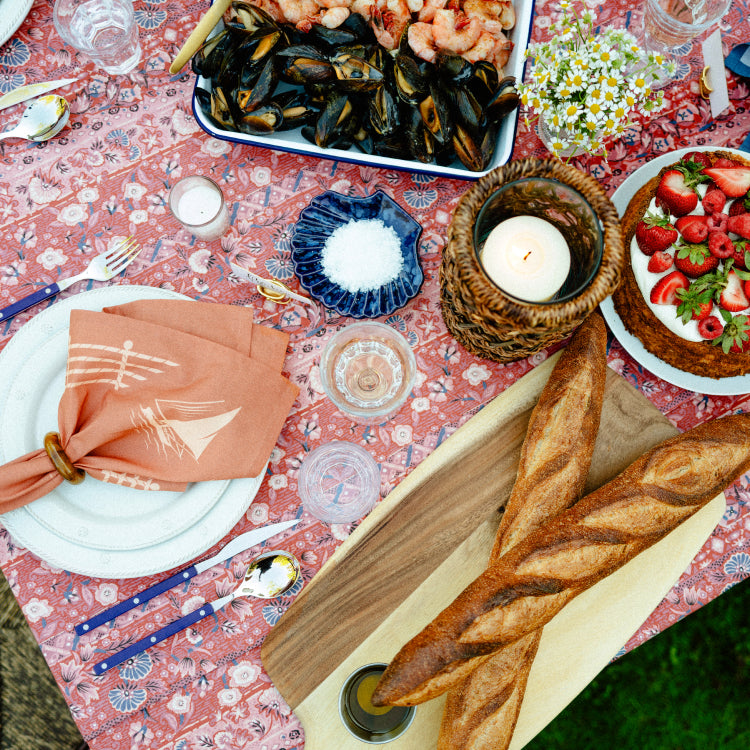 A summer table set with a red tablecloth, blue cutlery, and an orange sailboat napkin. 