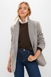 A woman models a Tuckernuck oversized gray herringbone blazer with suede elbow patches, layered over a brown sweater and white collared shirt, paired with blue jeans, standing against a plain white background.