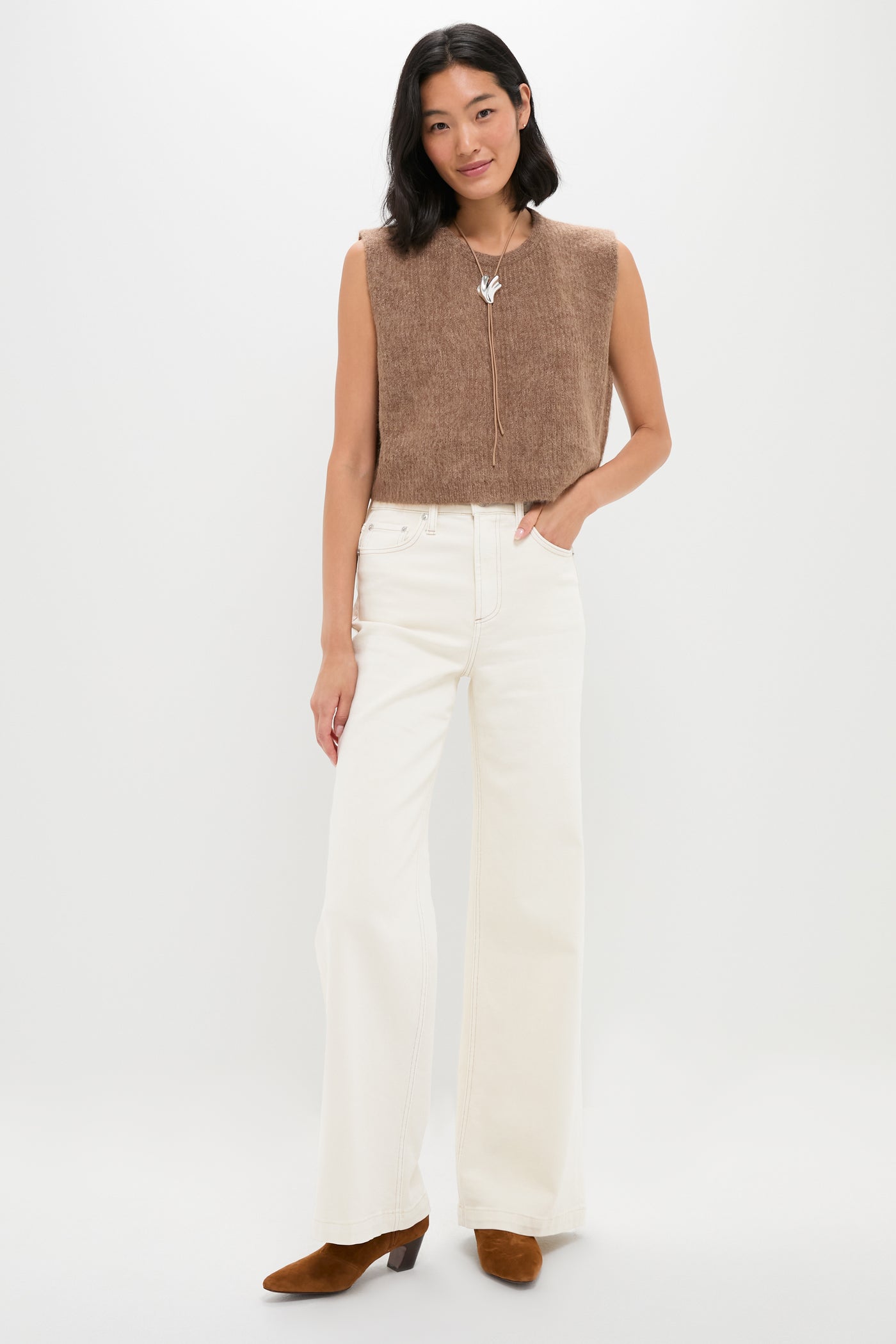 A woman poses against a white background wearing THE GREAT. sleeveless baby alpaca sweater vest with a boxy fit, paired with white high-waisted wide-leg pants, brown heeled boots, and a bird-shaped necklace.