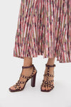 Cognac Studded Cage Heels with classic T-strap design, dark burgundy tone, and gold studs, paired effortlessly with a pleated multicolored striped midi skirt.