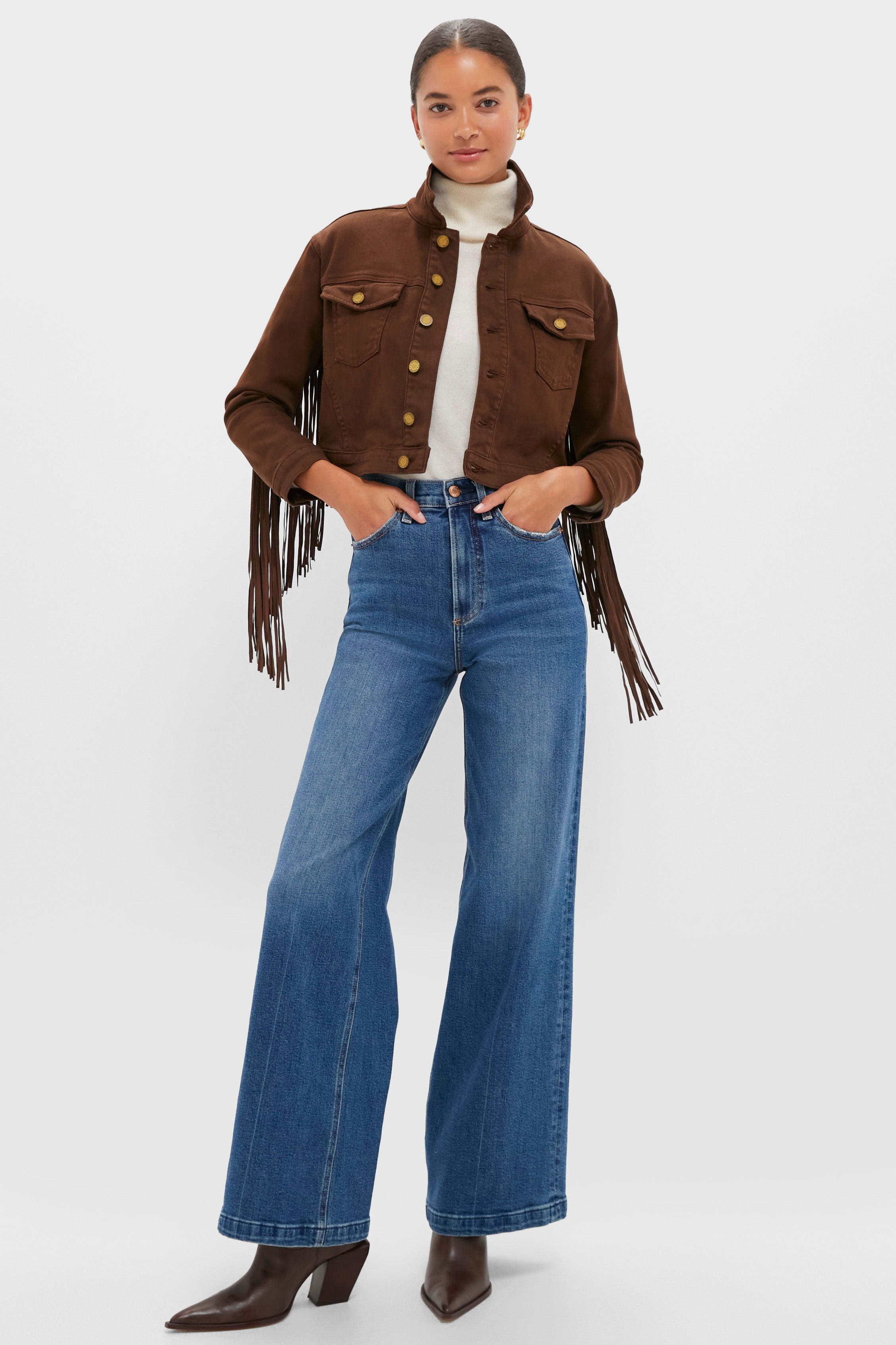 A woman in a L’AGENCE brown fringe denim jacket, white turtleneck, blue high-waisted wide-leg jeans, and brown pointed boots stands against a plain background.