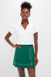 A woman stands wearing a white Tnuck Sport Short Sleeve Caroline Polo with SPF protection and a green skirt featuring decorative stitching near the hem.