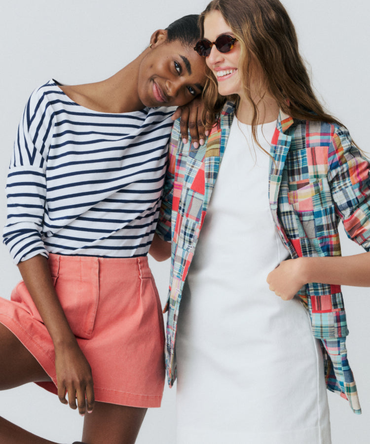 Two women standing in front of a white backdrop. One to the left in a blue and white striped tee and red shorts. One to the right in a madras blazer and white shift dress. 
