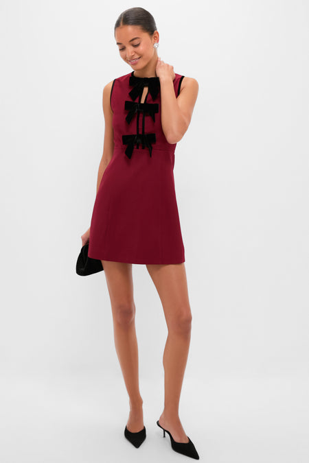 Model in Cranberry Velvet Bow Jaclyn Mini Dress, featuring classic black velvet bows, holding a black clutch, embodying an effortless, fun, and high-end style with elegant accessories.