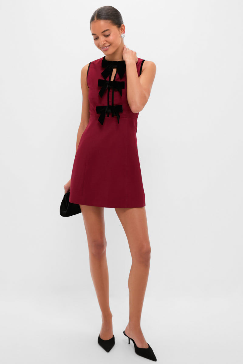 Model in Cranberry Velvet Bow Jaclyn Mini Dress, featuring classic black velvet bows, holding a black clutch, embodying an effortless, fun, and high-end style with elegant accessories.
