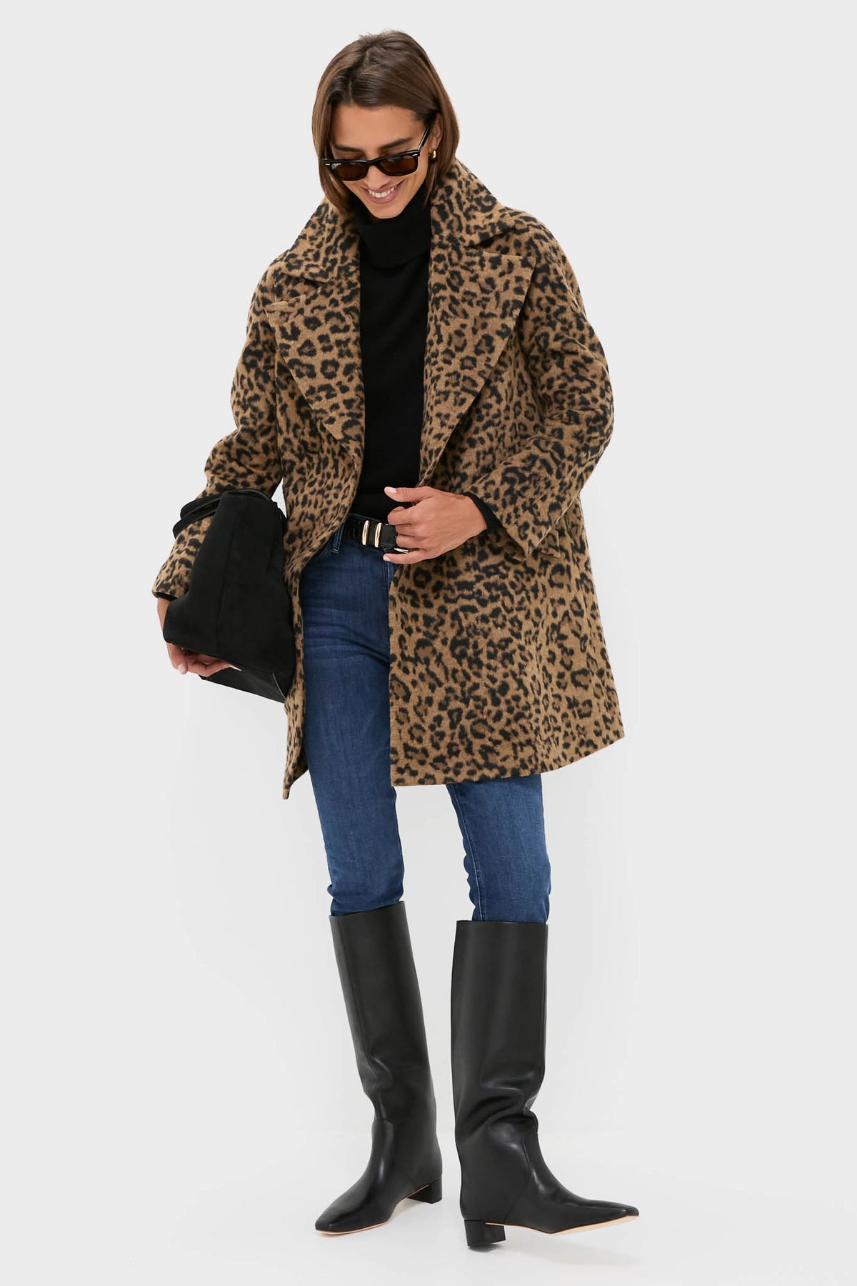 A person in a Tuckernuck double-breasted leopard-print coat, black turtleneck, blue jeans, knee-high black boots, and sunglasses holds a black bag against a plain background.