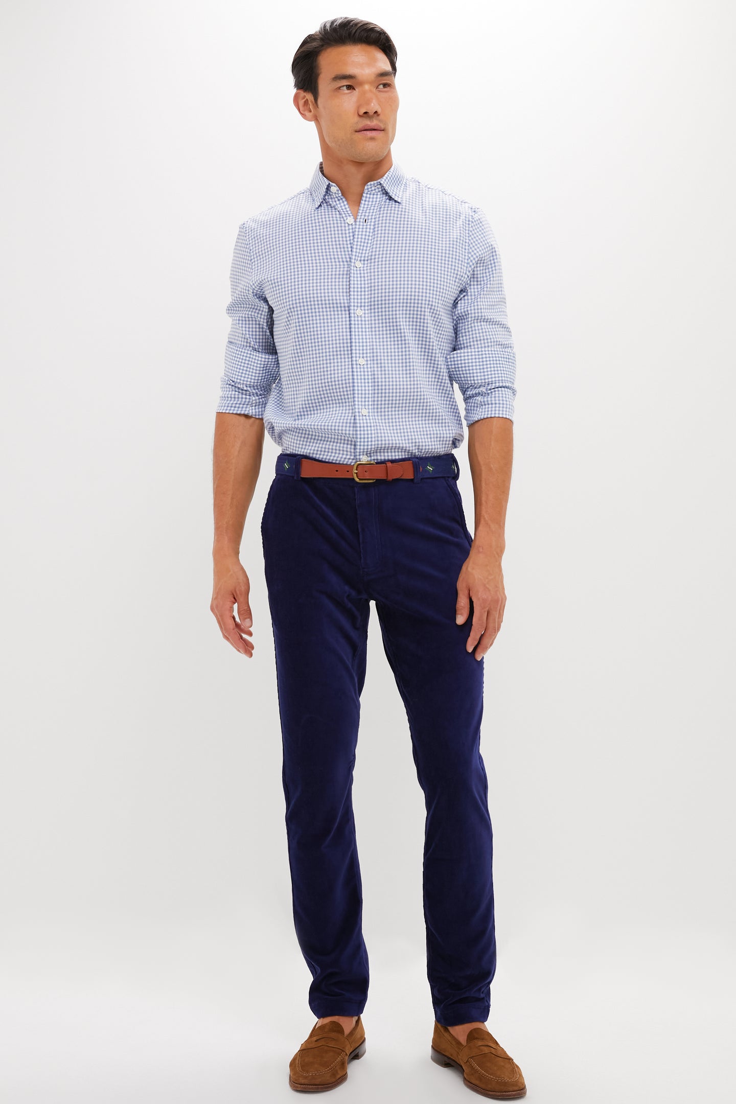 A man wears a light blue checkered shirt, blue REDVANLY performance corduroy trousers, a brown belt, and brown loafers against a plain white background.