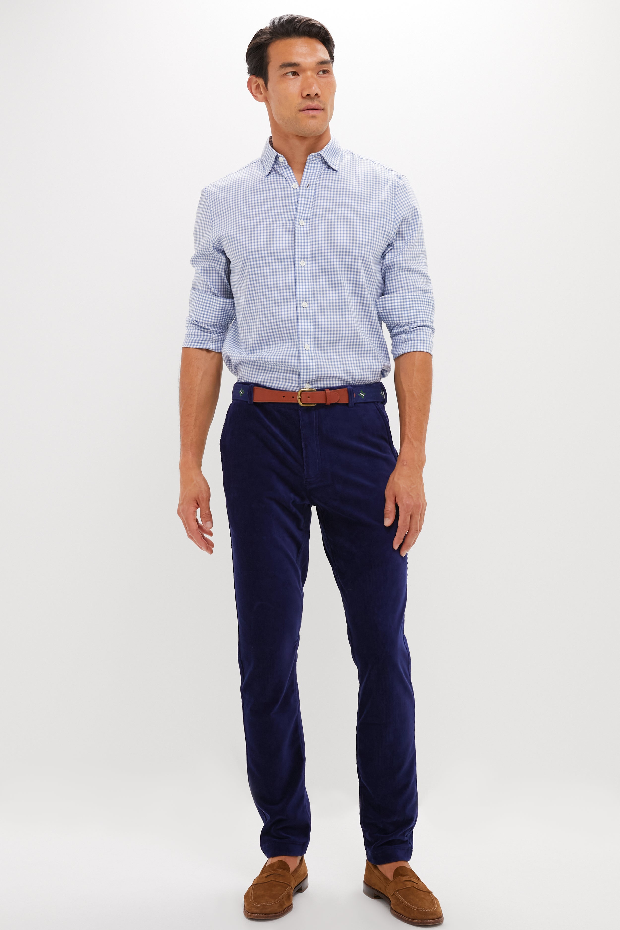 A man wears a light blue checkered shirt, blue REDVANLY performance corduroy trousers, a brown belt, and brown loafers against a plain white background.