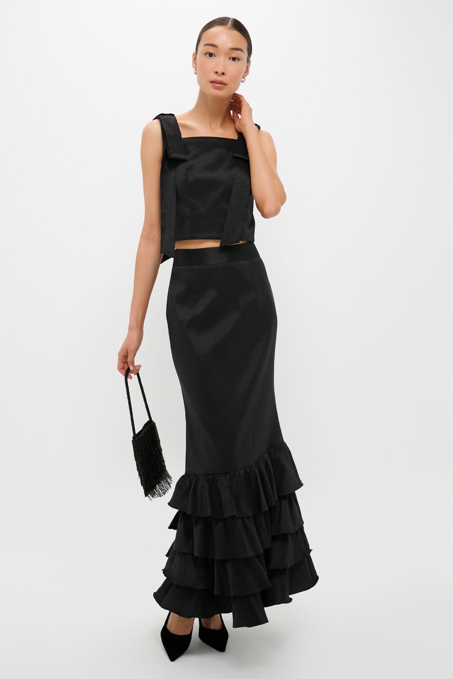 A woman wearing Tuckernuck's black sleeveless top and long black ruffle-hem maxi skirt holds a small black fringe bag while standing against a plain white background.