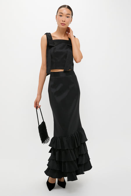 A woman wearing Tuckernuck's black sleeveless top and long black ruffle-hem maxi skirt holds a small black fringe bag while standing against a plain white background.