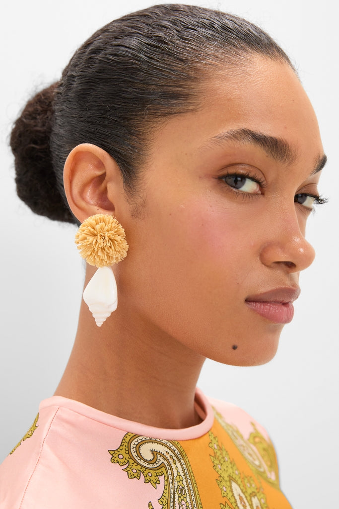 Close-up of a woman wearing Shell and Pom Elaine Earrings, featuring a straw-like pom at the lobe and a dangling seashell, embodying classic, effortless high-end style.