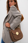 Woman wearing a light gray blazer and jeans carries the Taupe Agave Shoulder Bag in rich brown suede, showcasing its classic, effortless, high-end design with gold-tone hardware.