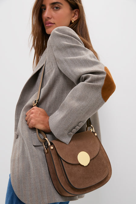 Woman wearing a light gray blazer and jeans carries the Taupe Agave Shoulder Bag in rich brown suede, showcasing its classic, effortless, high-end design with gold-tone hardware.