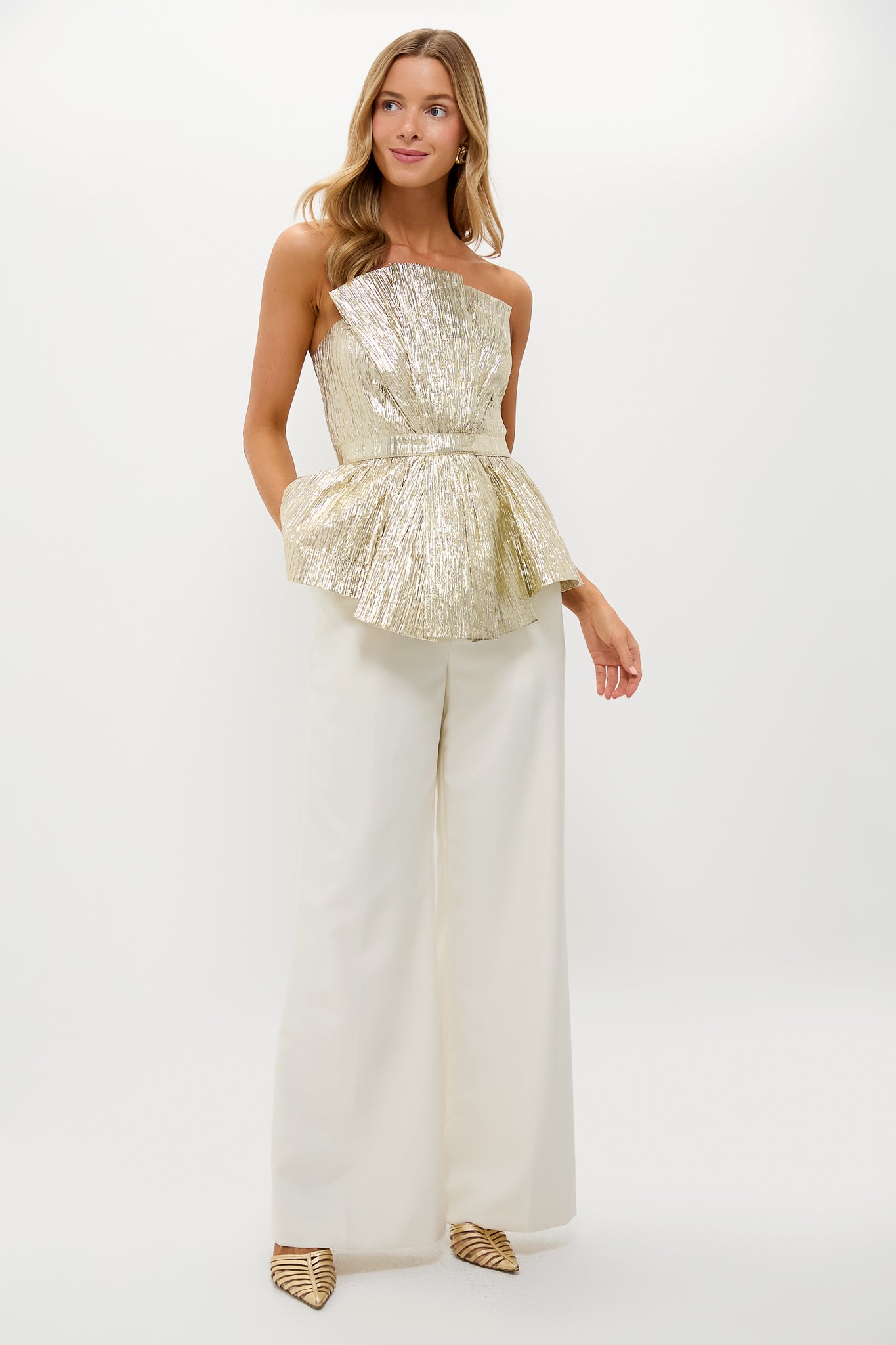 Woman models the Gold Plisse Iris Strapless Top with sculptural peplum, paired with ivory wide-leg trousers. Effortless, classic, and high-end party-ready look.