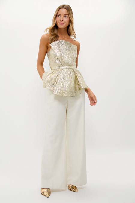 Woman models the Gold Plisse Iris Strapless Top with sculptural peplum, paired with ivory wide-leg trousers. Effortless, classic, and high-end party-ready look.