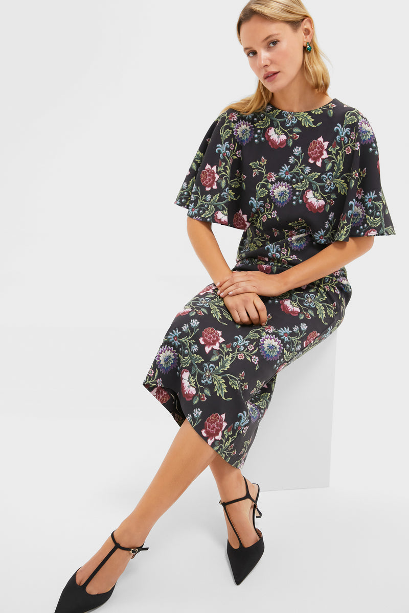 A woman sits on a white block, wearing a form-fitting Tuckernuck party dress with colorful floral patterns and black heels, set against a plain white background.