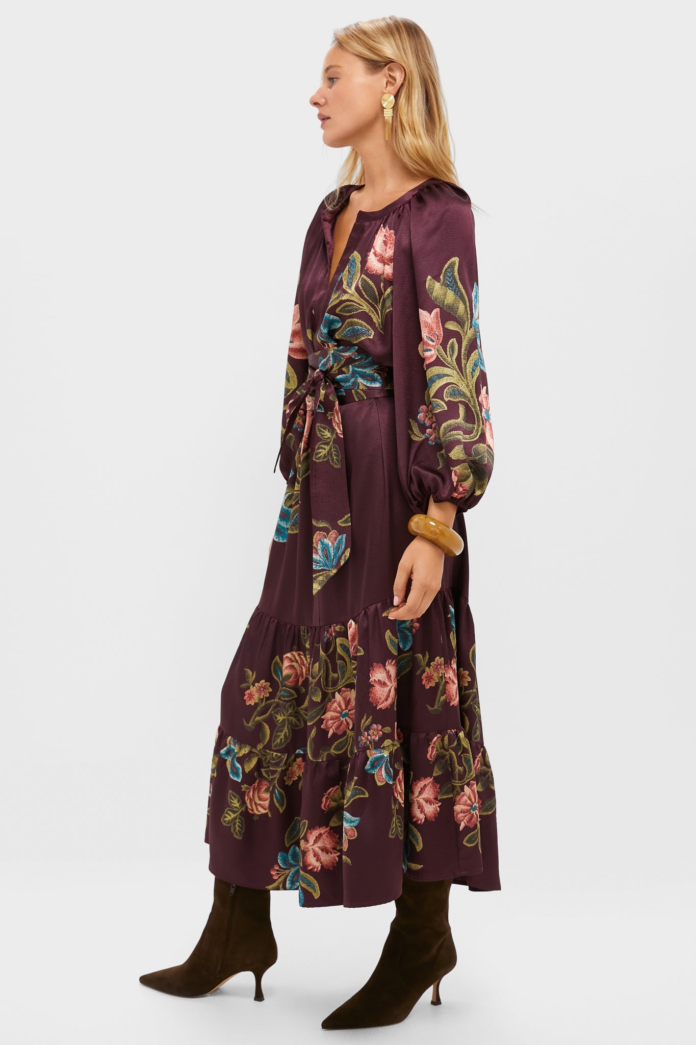 A woman in profile wears a Tuckernuck long-sleeve purple floral maxi dress with puffed sleeves and a removable waist belt, paired with brown heeled boots, against a plain background.