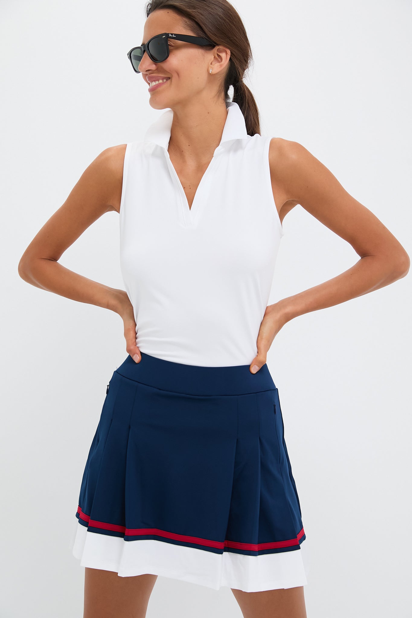 A woman stands confidently with hands on hips, wearing Tnuck Sport sunglasses, a white sleeveless Caroline polo, and a navy blue pleated skirt featuring red and white stripes.