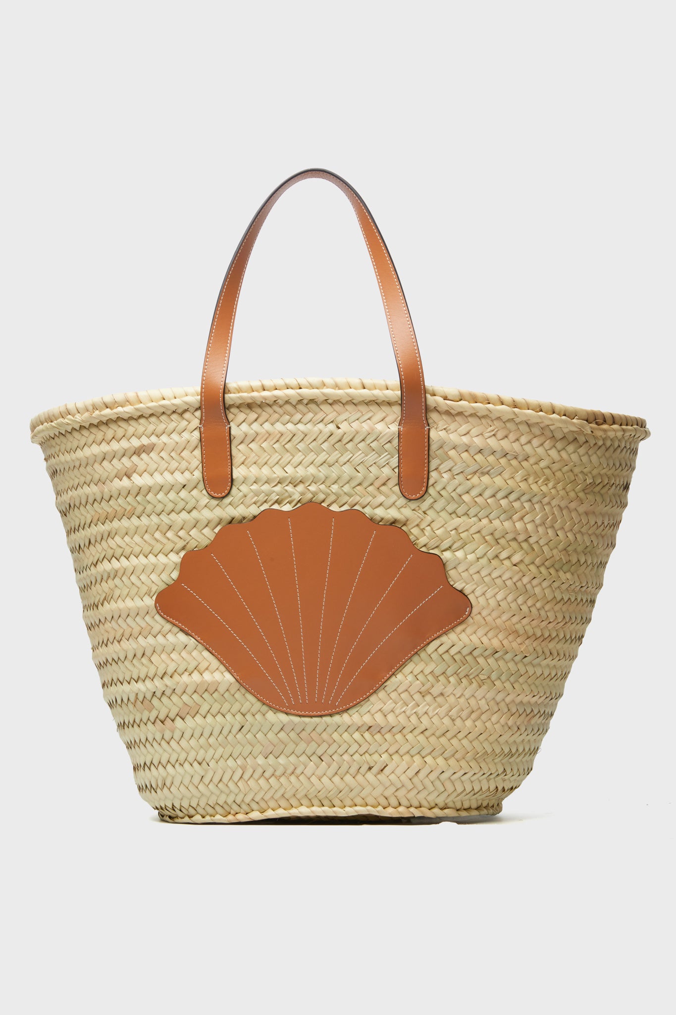 The Poolside handcrafted straw tote features brown leather handles and a shell-shaped leather patch on the front, making it a stylish beach bag.