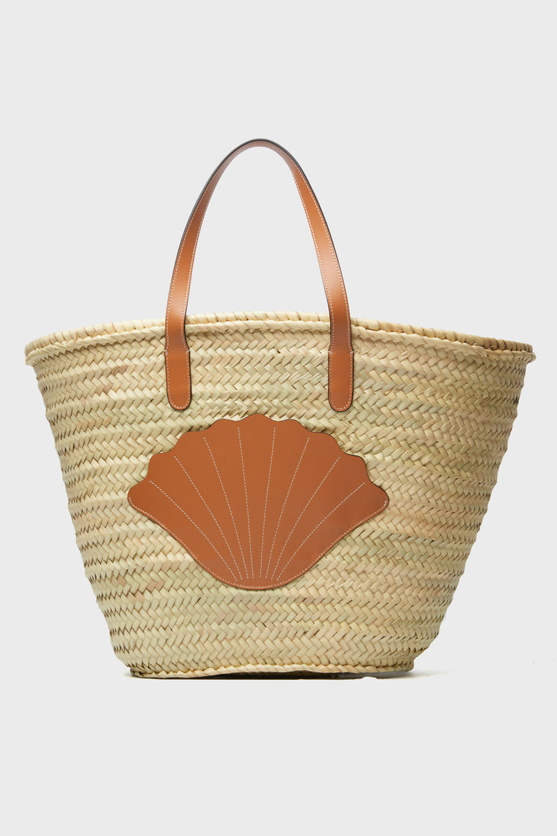 The Poolside handcrafted straw tote features brown leather handles and a shell-shaped leather patch on the front, making it a stylish beach bag.
