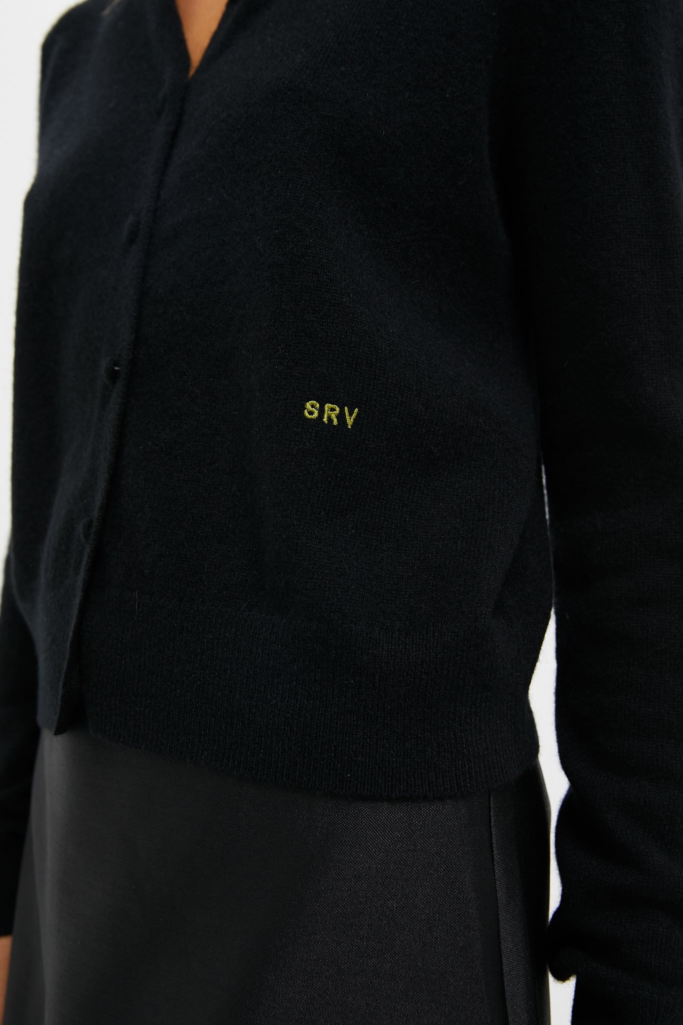 A person wearing a Tuckernuck black cashmere cardigan embroidered with SRV over a black skirt.