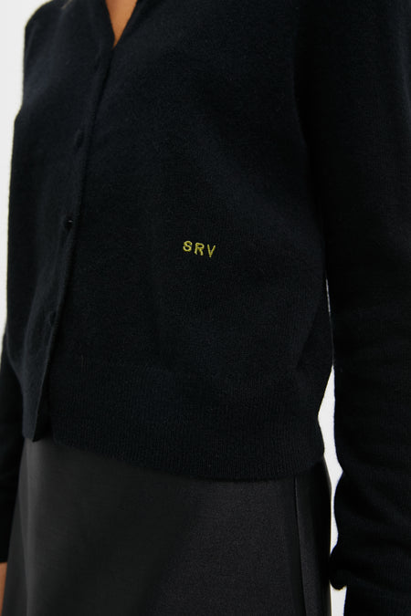 A person wearing a Tuckernuck black cashmere cardigan embroidered with SRV over a black skirt.