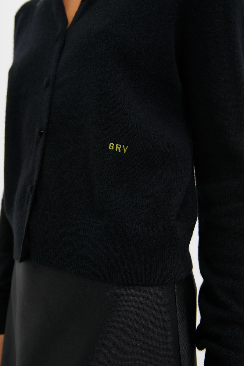 A person wearing a Tuckernuck black cashmere cardigan embroidered with SRV over a black skirt.