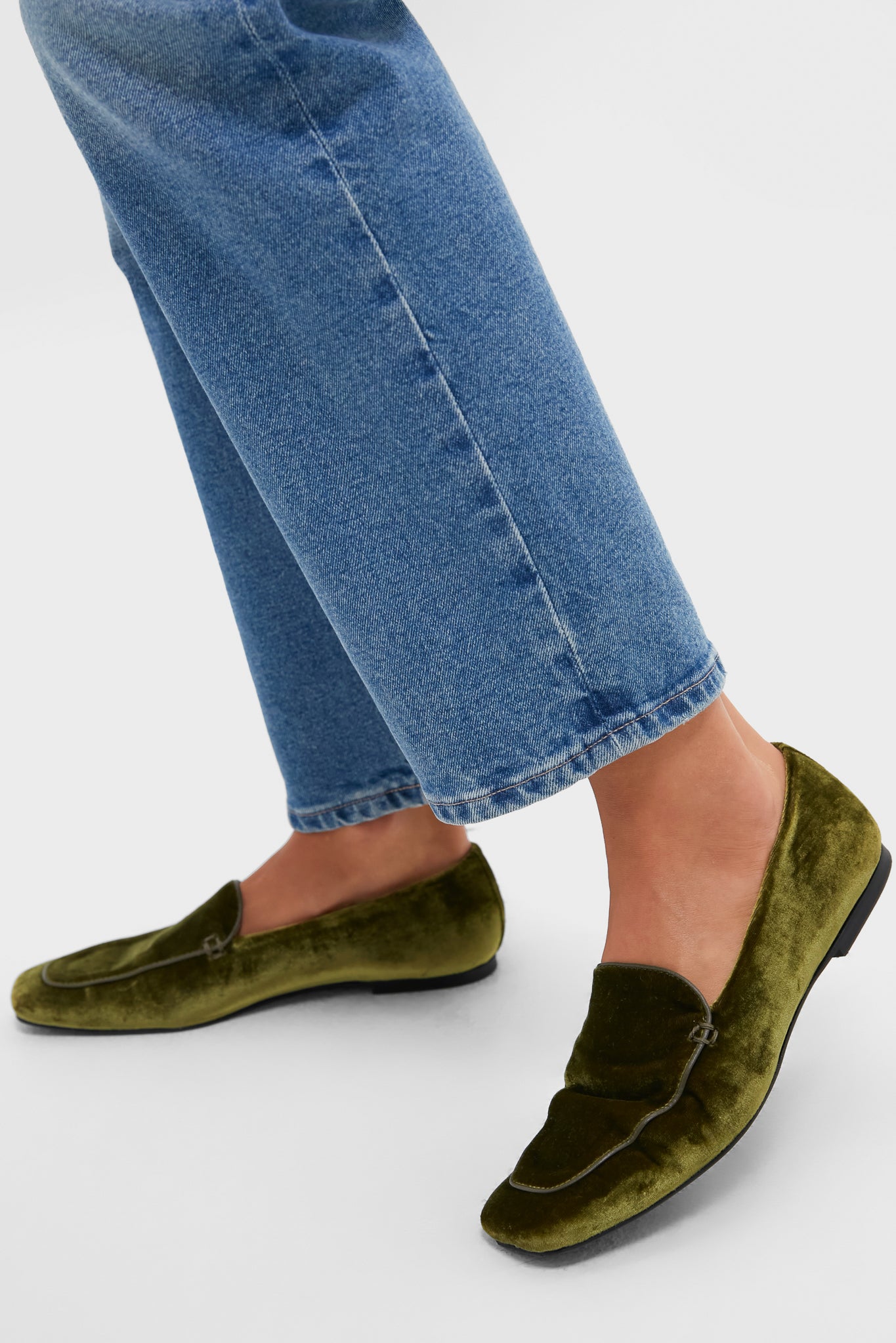 A person stands on a white background wearing blue jeans and green velvet loafers with sleek leather trim from Tuckernuck Shoes.
