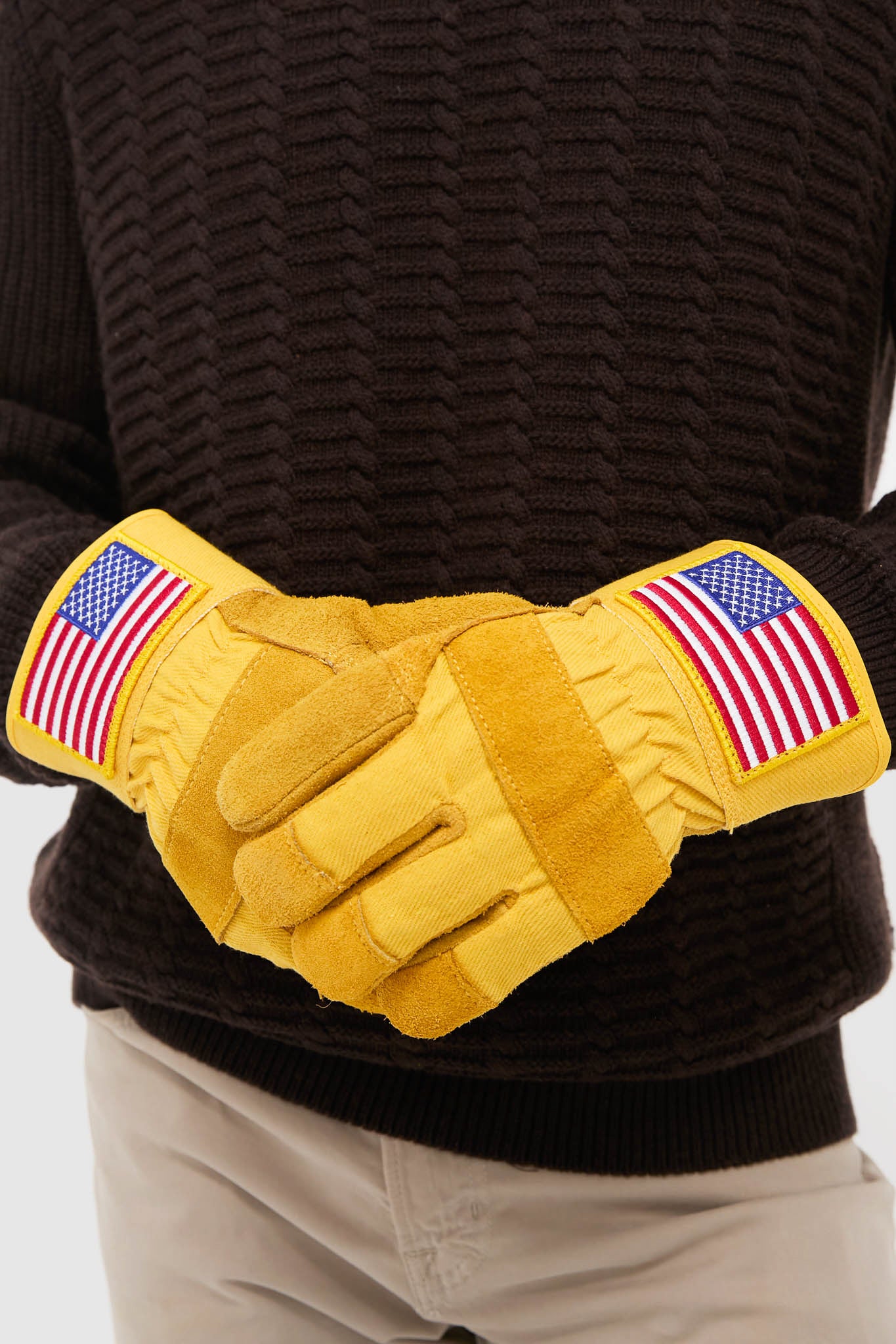 A person wearing Brisket Supply insulated work gloves with American flag patches stands with hands clasped, dressed in a black sweater and beige pants.