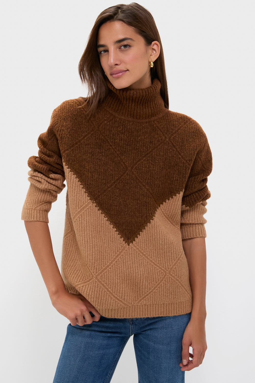 A person models a Hyacinth House color block sweater with geometric patterns, midweight knit turtleneck, and blue jeans in a simple light setting.