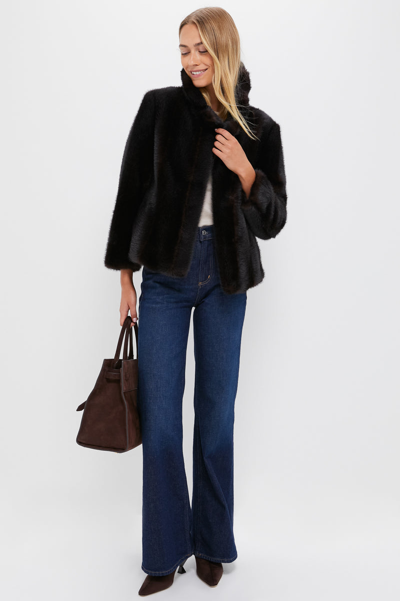 A woman in a Tuckernuck black faux fur coat, white top, blue wide-leg jeans, and brown heels holds a dark brown handbag against a plain white background—an effortlessly chic winter statement look.