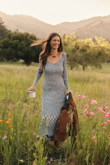 Woman in a blue sweater dress walking through a field of flowers holding a coffee mug and a brown jacket. 