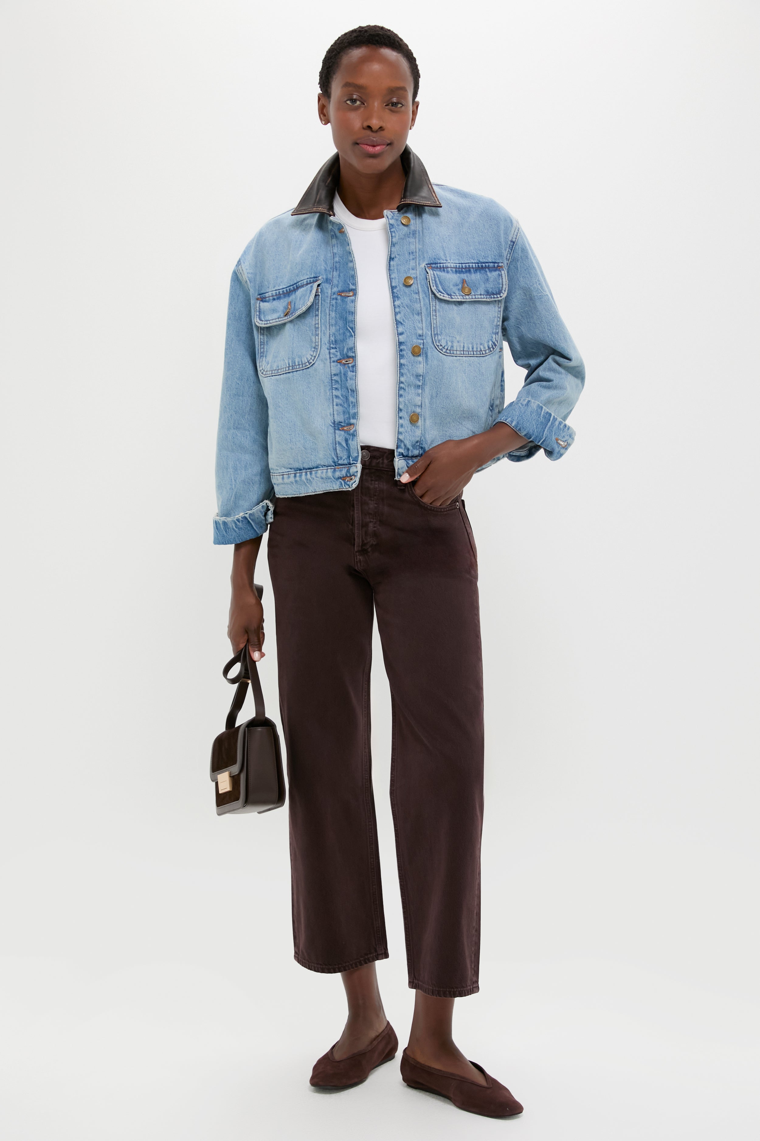 A woman stands against a white background wearing a SIMKHAI cropped denim jacket with a contrast collar, styled with a white shirt, brown cropped pants, brown flats, and a small handbag.