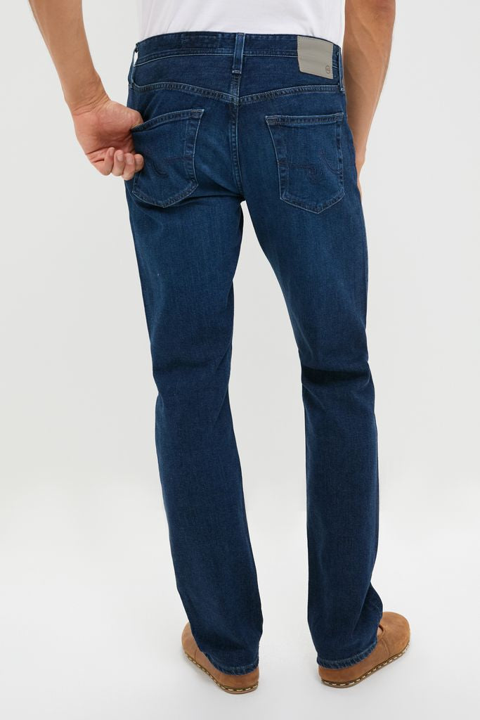 Back view of The Graduate in Crusade Wash Denim (34 Inseam) by AG Jeans, showcasing classic, effortless style with visible back pockets and leather-look patch, paired with casual shoes.