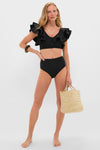 Model showcases the Black Mila Two Piece swimsuit with ruffled cap sleeves and high-waisted bottoms, accessorized with chunky green earrings and a woven straw tote, embodying effortless, high-end style.