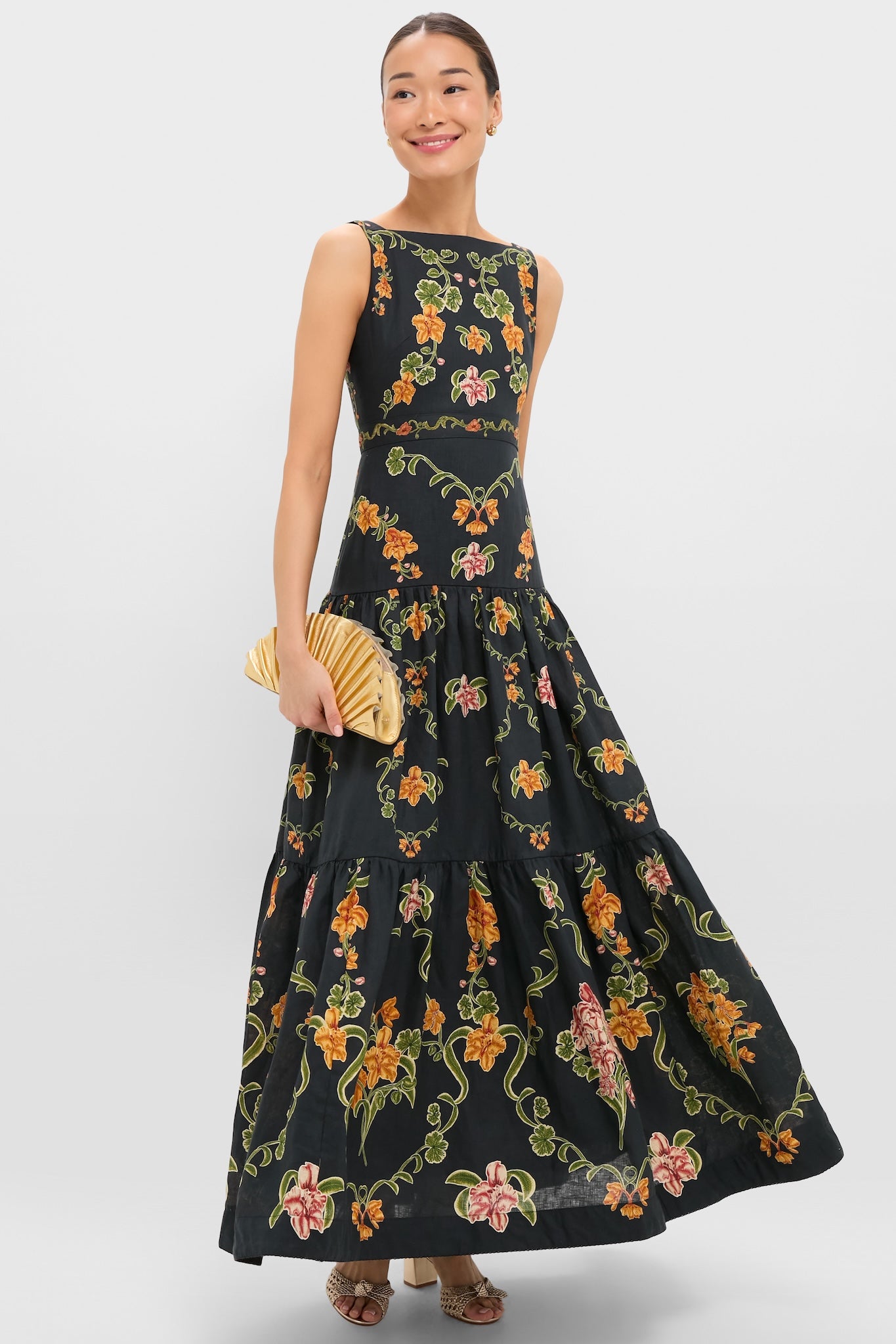 Woman elegantly showcases the Gold Nala Mini Clutch, a classic, high-end pleated bag. Effortlessly pairs with her floral maxi dress and open-toe heels for a fun, stylish look.