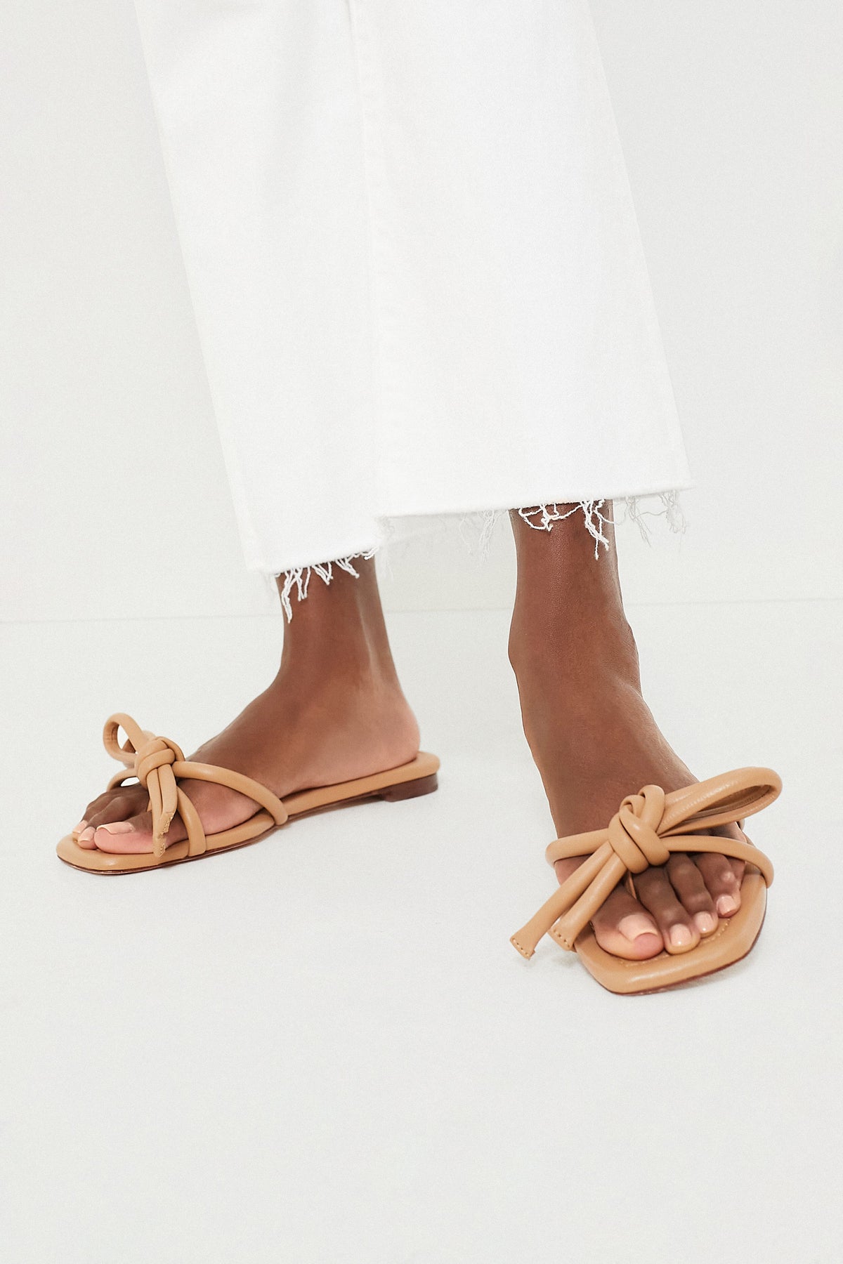 Dune Nappa Hadley Leather Bow Flat Sandal: Classic, effortless nude-tan slides with knotted straps, showcased with cropped white denim, embodying a fun, high-end style.