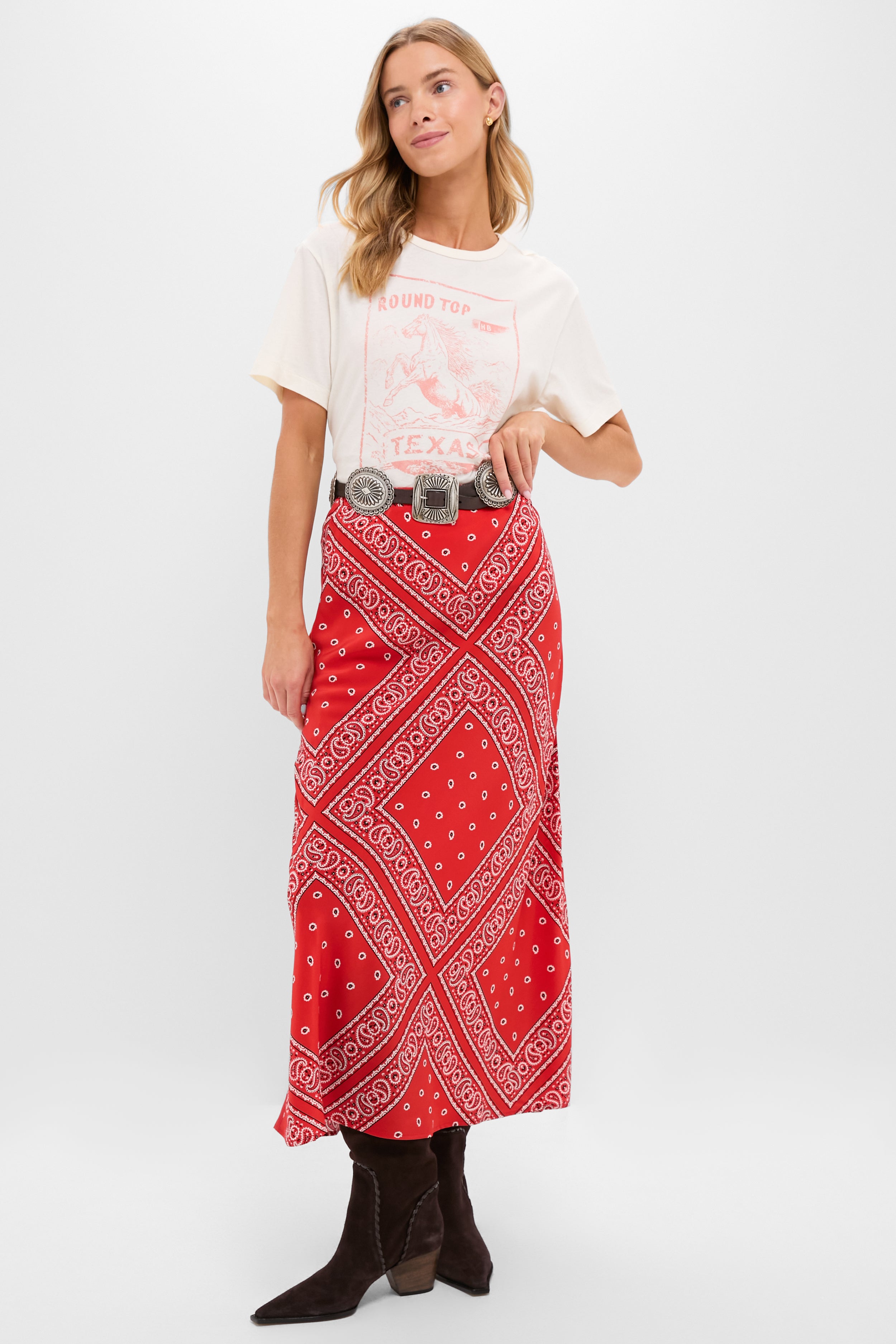 A woman wears a Hunter Bell vintage-inspired Western tee, red bandana-print maxi skirt, black western belt, and brown pointed-toe boots against a plain white background.