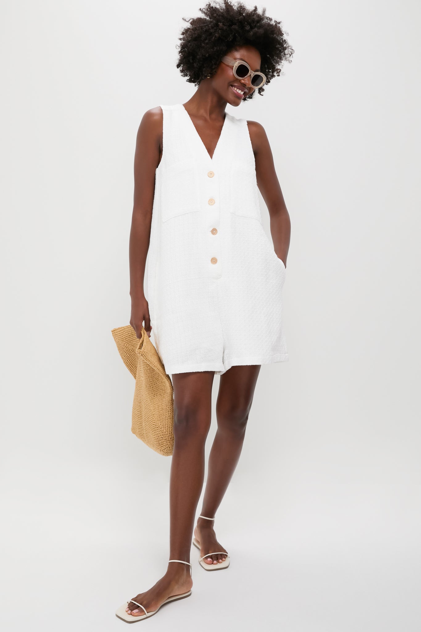 A person wears a sleeveless white Tuckernuck Myla Romper with large buttons and textured fabric, accessorized with a straw bag, sunglasses, and sandals.
