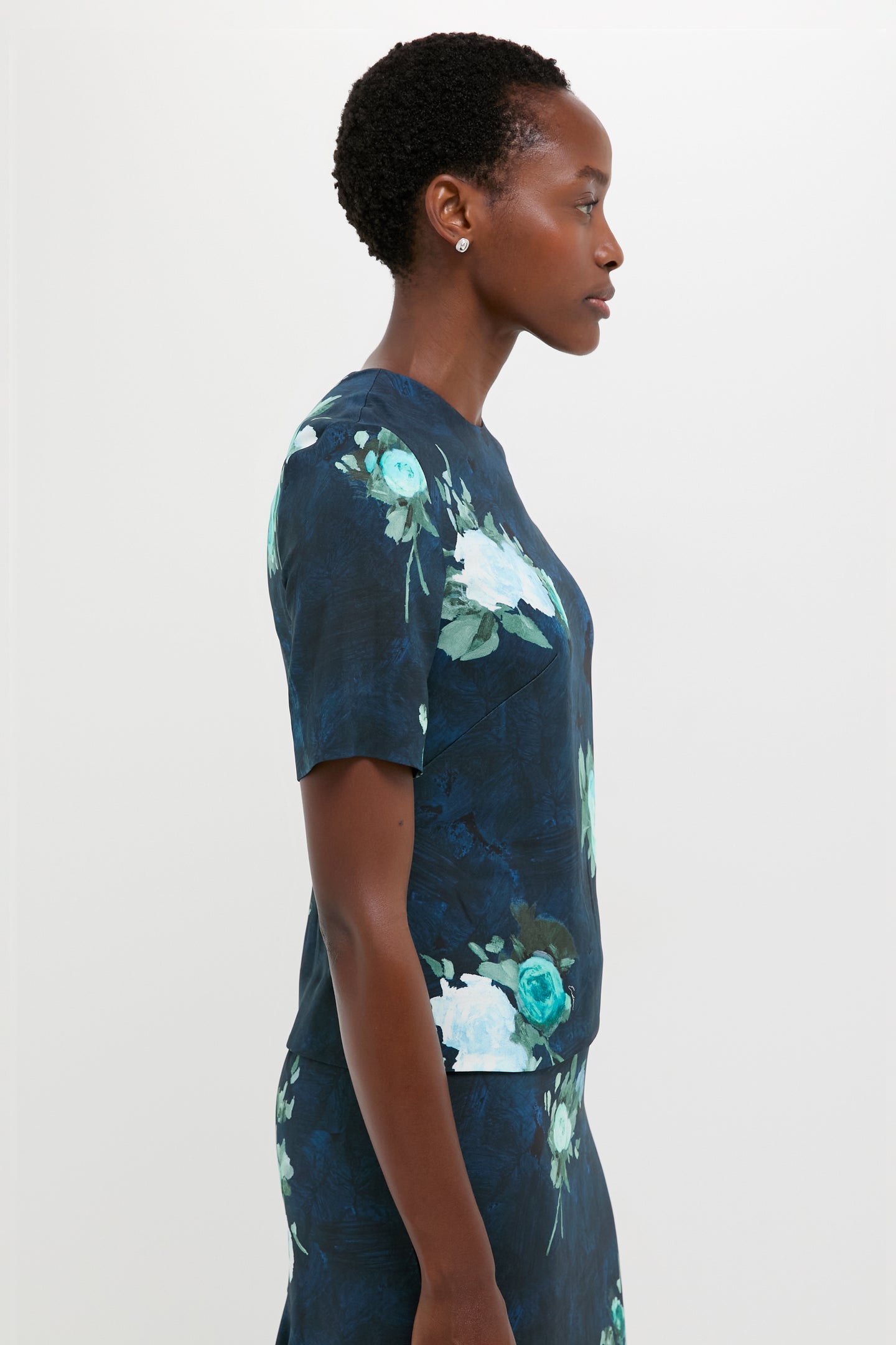A woman in profile wears an ERDEM navy blouse and skirt with a green and white abstract rose print, set against a plain light background.